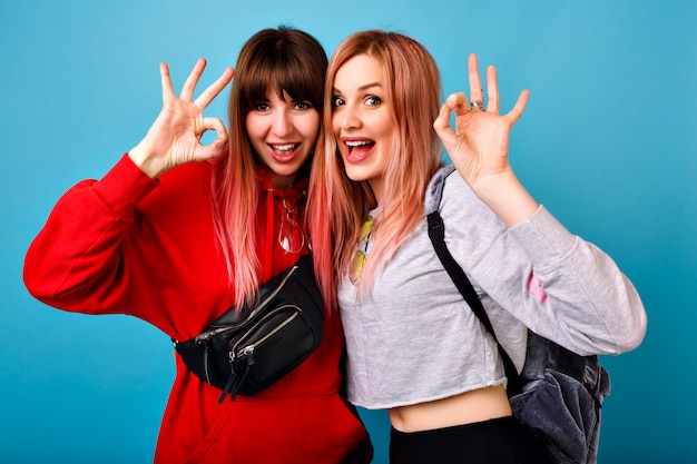 Two pretty young hipster woman showing ok gesture, lifestyle portrait, smiling and looking, happy couple of friends, casual bright sportive outfits.