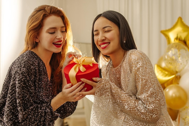 Two pretty young interracial ladies in dresses happily unpack red gift box indoors Surprise concept