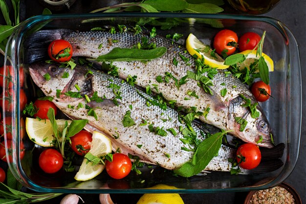 Two raw seabass in a baking dish with spices on an black table. Top view