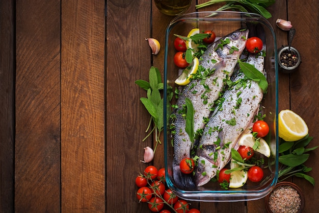 Two raw seabass in a baking dish with spices on an old wooden table. Top view