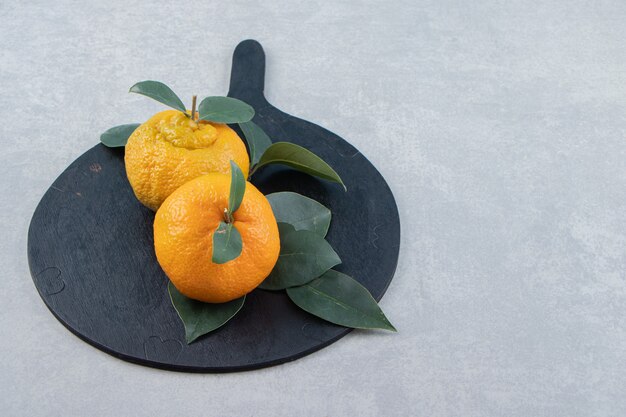 Two ripe tangerines with leaves on black cutting board. 