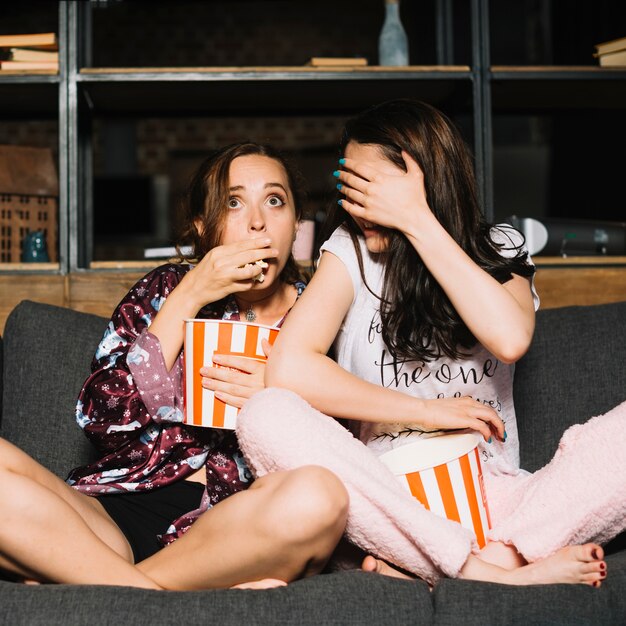 Two scared female friends eating popcorn while watching television