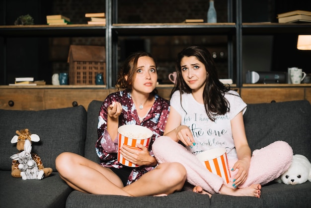 Two scared women sitting on sofa with popcorn watching television