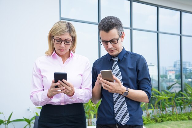 Two serious business colleagues using phones with city building glass 