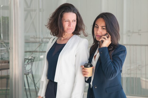 Two serious business women calling on phone and drinking coffee