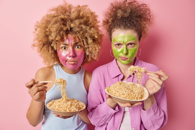 Two serious curly haired women eat noodles have strict displeased expressions apply beauty mask on face for skin treatment dressed in casual clothes stand next to each other isolated on pink wall