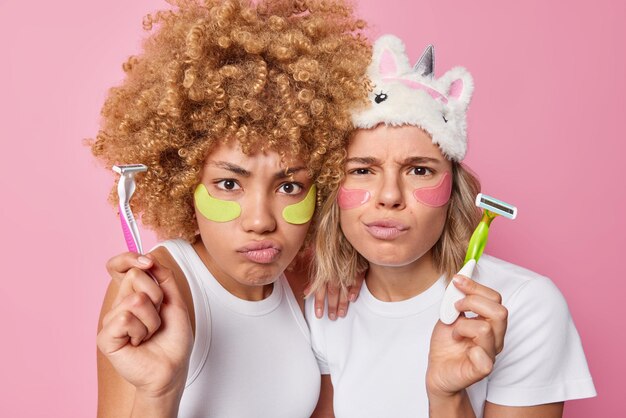 Две серьезные женщины хмурятся с мрачным выражением лица, наносят гидрогелевые пластыри под глаза, чтобы увлажнить нежную кожу, держат бритвы для бритья, одетые в повседневные футболки, изолированные на розовой стене