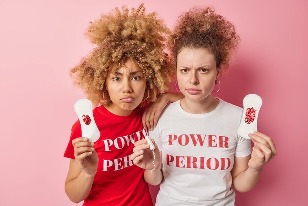 Two serious young women with curly hair hold intimate hygiene products give advice about women health and menstruation dressed in casual t shirts isolated over pink background Absorbency items