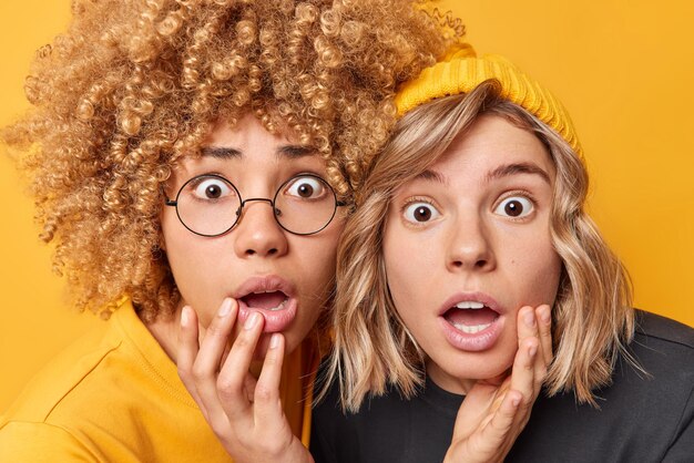 Two shocked young women stare at camera with frightened facial expressions hold breath cannot believe in something amazing dressed in casual clothes isolated over yellow background Omg concept