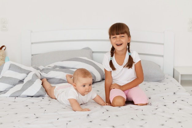 Two sibling playing at home, posing on bed, elder child looking smiling directly at camera, dark haired elder kid wearing casual style playing with toddler child lying on tummy.