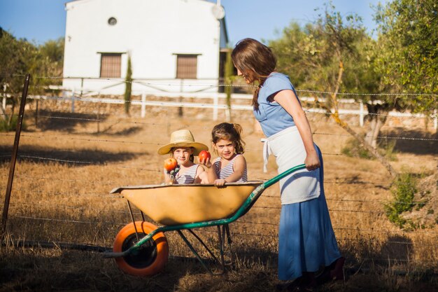 Two sister holding red apple sitting in wheelbarrow being pushed by woman