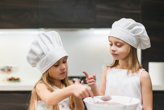Two sisters in kitchen wearing chef's hat looking at messy finger while taking cocoa powder