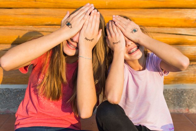 Two sisters sitting outside covering their eyes with eyes tattoo on palm