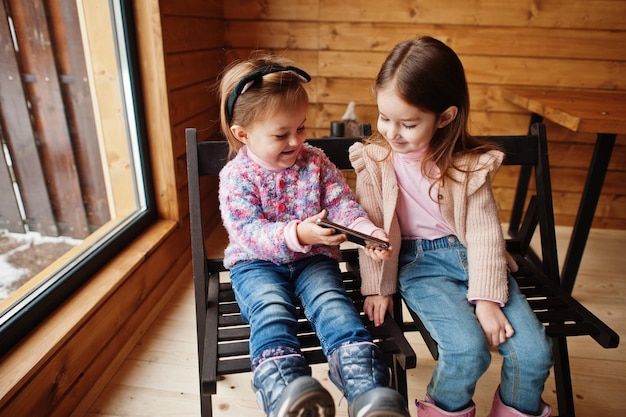 Two sisters watching at mobile phone in modern wooden house sitting on chair