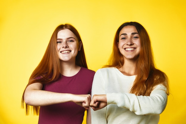 Two smiled redhead caucasian girls are standing shoulder to shoulder and their fists are together, on the yellow dressed in casual clothes