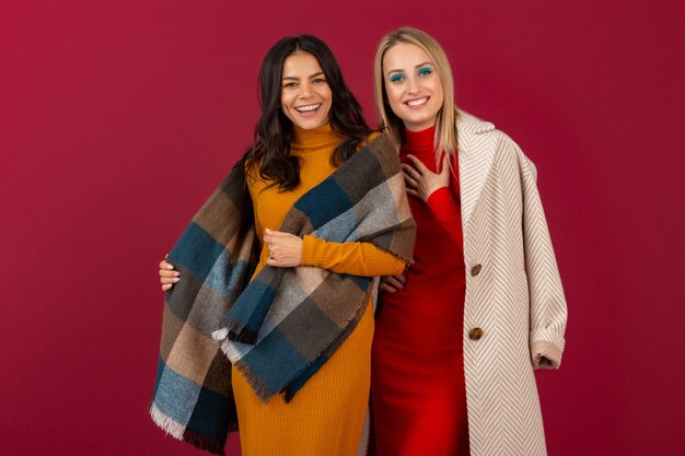 Две улыбающиеся привлекательные стильные женщины в осенне-зимнем модном платье и пальто позируют изолированно на красной стене