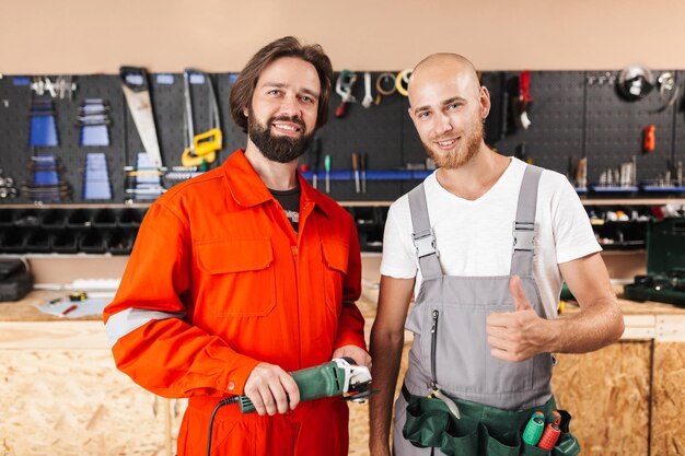 Two smiling builders in work clothes happily looking in camera spending time in workshop with variety of tools on background