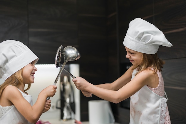 Two smiling cute sisters in chef's hats are fighting with kitchen utensil