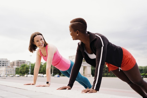 Две улыбающиеся разноплановые молодые женщины в спортивной одежде делают упражнения на доске, вместе тренируясь на городской площади