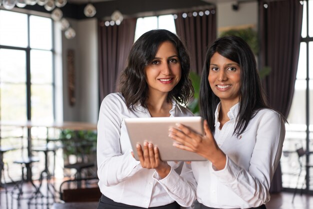 Две улыбающиеся женщины-коллеги, используя планшетный компьютер в кафе.