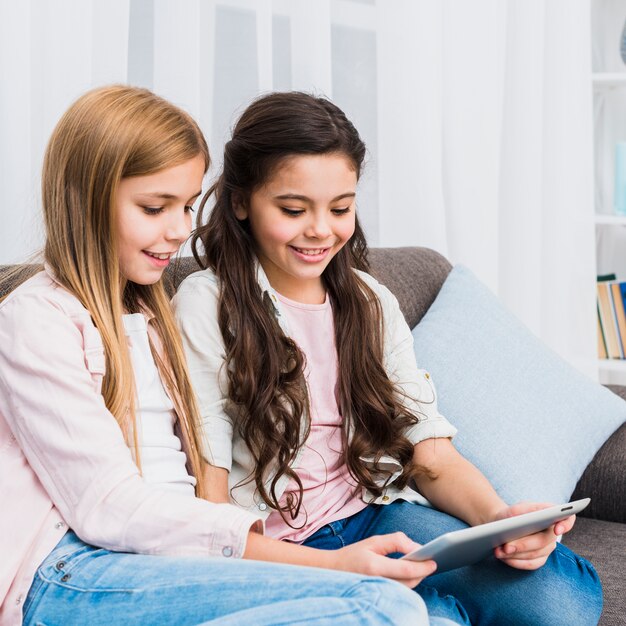 Two smiling girls sitting on sofa looking at digital tablet