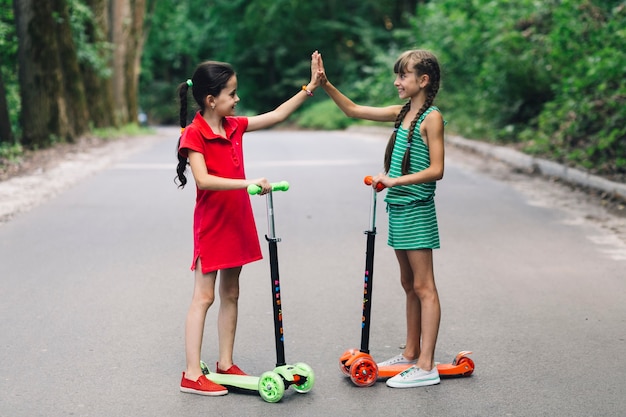 Two smiling girls standing on scooter giving high five gesture on road