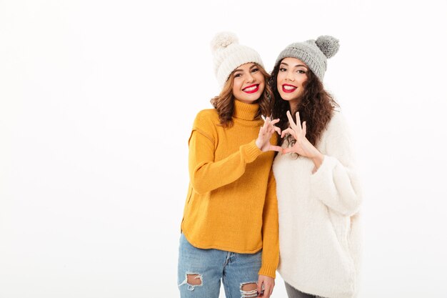 Two smiling girls in sweaters and hats making heart sign  over white wall