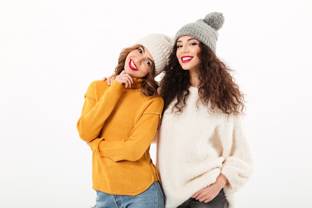Two smiling girls in sweaters and hats posing together  over white wall