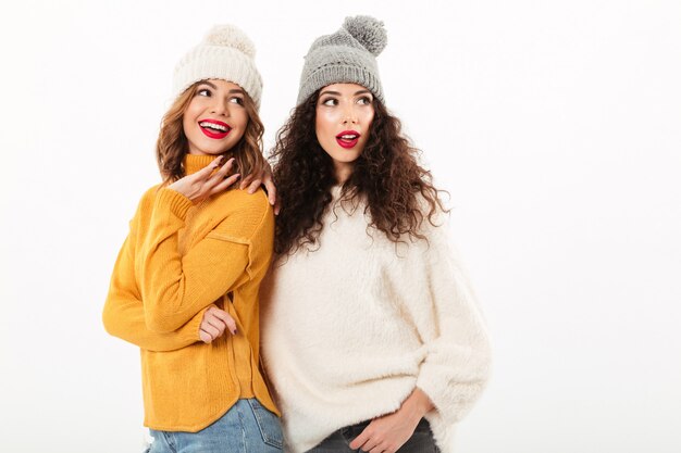 Two smiling girls in sweaters and hats standing together while looking away over white wall
