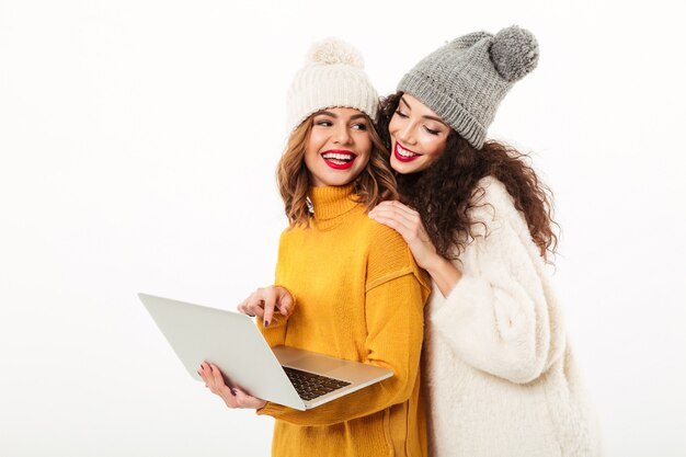 Due ragazze sorridenti in maglioni e cappelli che stanno insieme mentre per mezzo del computer portatile sopra la parete bianca