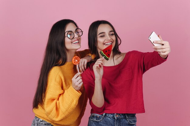 Two smiling girls take selfie on their phones posing with lollipops