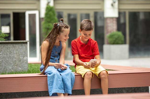 Two smiling kids, boy and girl playing smartphone in town, city in summer day. Concept of childhood, happiness, sincere emotions, carefree lifestyle. Little caucasian models in bright clothes.