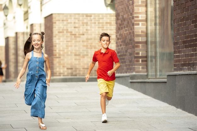 Two smiling kids, boy and girl running together in town, city in summer day