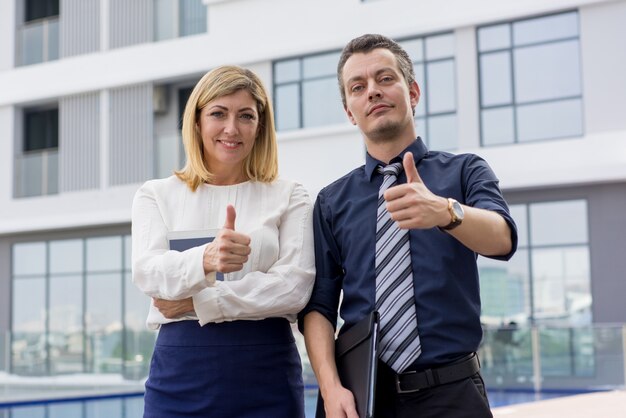 Two smiling male and female business people showing thumbs up outdoors.