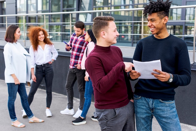 Two smiling man discussing about document with their friends standing at background