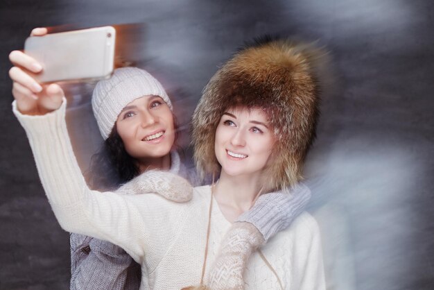 Две улыбающиеся женщины в зимних шапках фотографируют на смартфон.