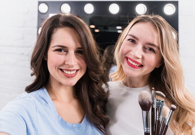 Две улыбающиеся женщины с кистями, делающие селфи на косметическое зеркало