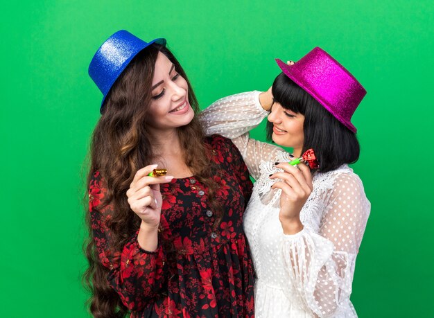 Две улыбающиеся молодые тусовщицы в шляпе для вечеринки держат в руках рожок, глядя друг на друга, одна кладет локоть на плечо подруги, а руку на свою голову, изолированную на зеленой стене