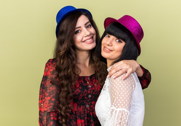 Two smiling young party womans wearing party hat one holding another one by shoulder both looking at front isolated on olive green wall
