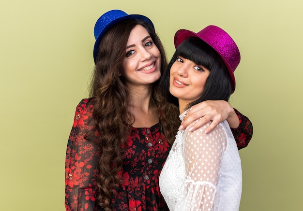 두 명의 웃고 있는 젊은 파티 여성이 올리브 녹색 벽에 격리된 정면을 바라보며 어깨를 잡고 다른 모자를 쓰고 파티 모자를 쓰고 있습니다.