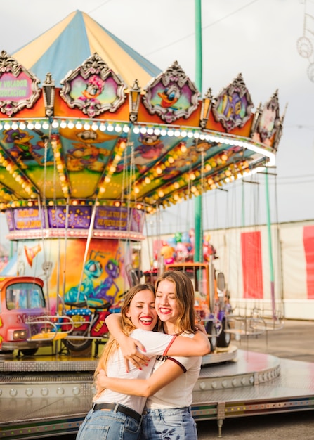 Two smiling young woman embracing in front of illuminated colorful carousel