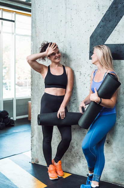 Two smiling young women with exercise mat in gym