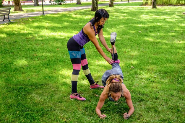 Две спортивные женщины разогреваются на лужайке в летнем парке.