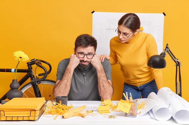 Free photo two students prepare for upcoming examination sit at office desk with papers blueprints and sketches. frustrated unhappy bearded man feels tired after preparing architectural project. teamworking