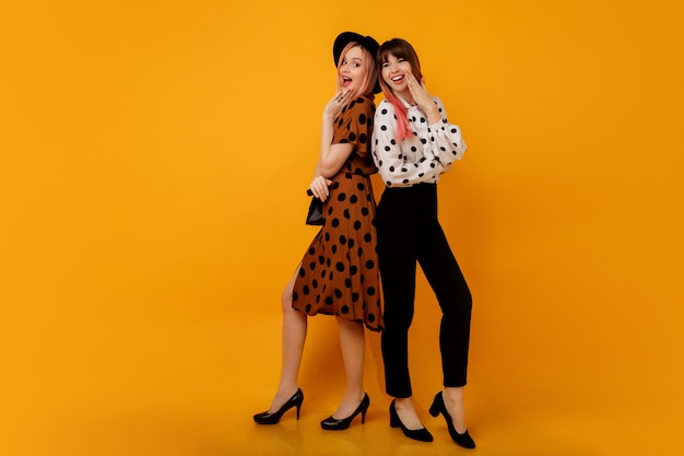 Free photo two stylish elegant women in dresses posing over yellow wall