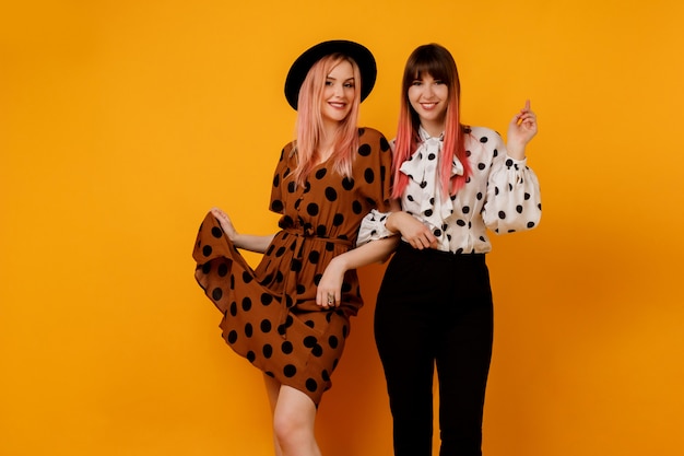 Free photo two stylish elegant women in dresses posing over yellow wall