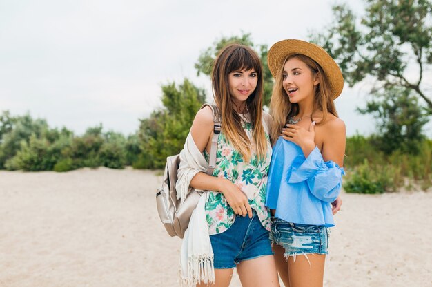 Две стильные симпатичные улыбающиеся женщины на летних каникулах на тропическом пляже, друзья путешествуют вместе