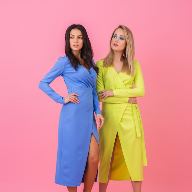 Две стильные сексуальные привлекательные женщины позируют в полный рост на розовой стене в стильных красочных платьях синего и желтого цвета, тренд летней моды