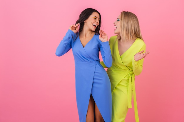 Two stylish smiling attractive women having fun posing on pink wall in stylish colorful dresses of blue and yellow color, spring fashion trend