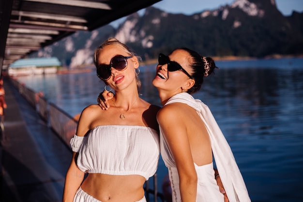 Two stylish woman in white summer clothes wearing sunglasses at dusk